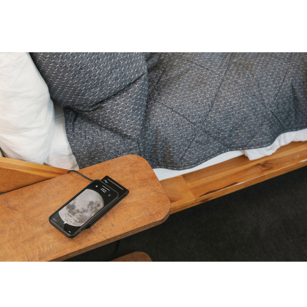 Peak-Design-Mobile-Wireless-Charging-Stand-5 Peak Design Mobile Wireless Charging Stand -mobiililatausteline pitää älypuhelinta, jossa näkyy kuun kuva puisella yöpöydällä, sinisen tikattua peittoa ja valkoista tyynyä vasten.