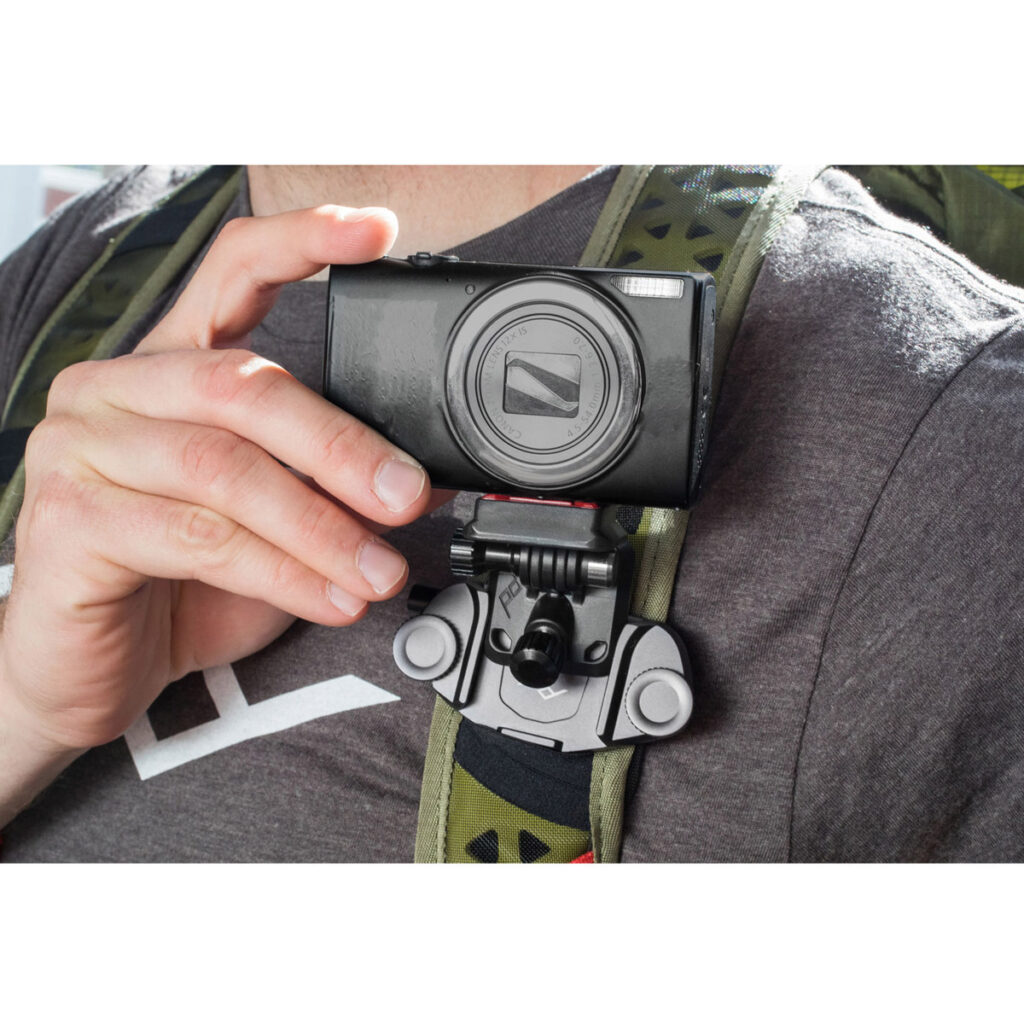 Peak-Design-POV-Kit4 Harmaaseen paitaan ja reppuun pukeutunut henkilö pitää mustaa kameraa, joka on kiinnitetty reppuhihnaan Peak Design POV Kit for Capture -laitteella.