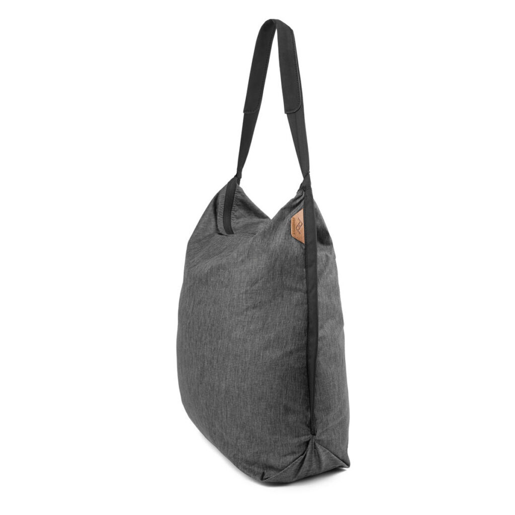 Peak-Design-Packable-Tote---Charcoal2 Peak Design Packable Tote in Charcoal -mallistossa on mustat olkahihnat ja ruskehtava nahkalappu edessä valkoista taustaa vasten.