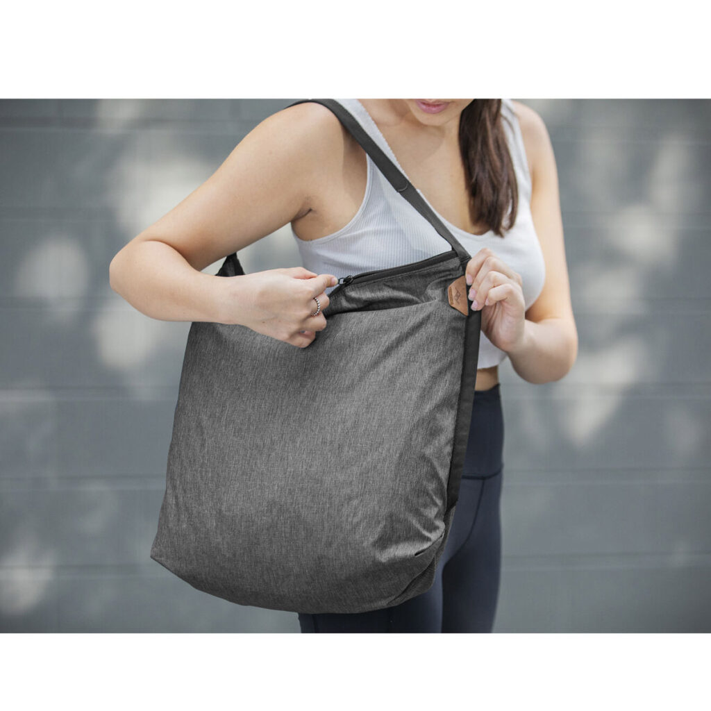 Peak-Design-Packable-Tote---Charcoal5 Urheiluvaatteet päällä oleva nainen pitelee Peak Design Packable Tote - Charcoal -laukkua toisella kädellä ja avaa sen vetoketjun toisella kädellä, ja taustalla on sumea harmaa tausta, joka korostaa laukkua ja hänen toimintaansa.