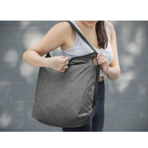 Peak-Design-Packable-Tote---Charcoal5 Urheiluvaatteet päällä oleva nainen pitelee Peak Design Packable Tote - Charcoal -laukkua toisella kädellä ja avaa sen vetoketjun toisella kädellä, ja taustalla on sumea harmaa tausta, joka korostaa laukkua ja hänen toimintaansa.