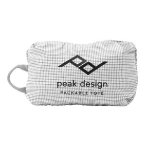 Peak-Design-Packable-Tote--Rawl3 Pieni, suorakulmainen, valkoinen Peak Design Packable Tote - Raw, jossa on ruudukkokuvio, sivukahva, musta "PACKABLE TOTE" -teksti ja Peak Designin logo edessä.