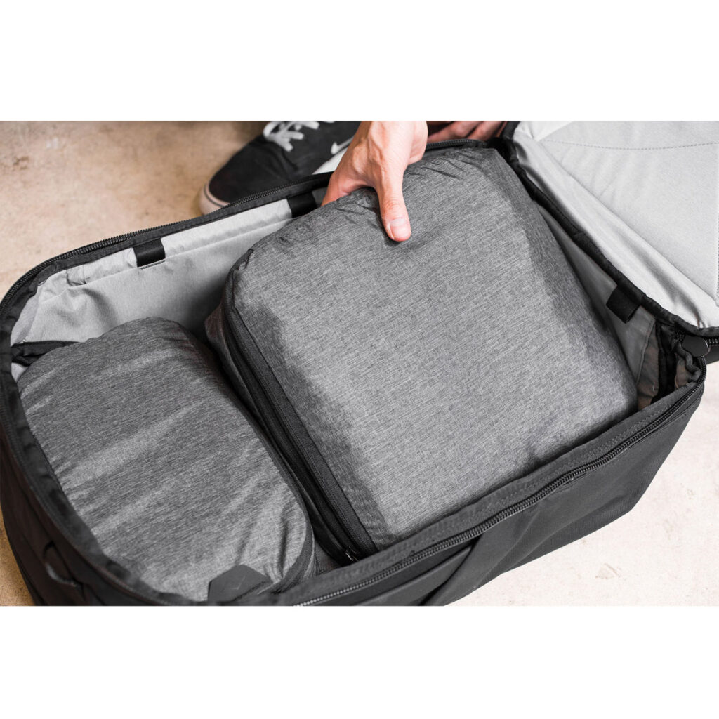 Peak-Design-Packing-Cube-medium--Charcoal6 Henkilö asettaa Peak Design Packing Cube Medium - Charcoal -kuution mustaan matkalaukkuun toisen kuution viereen, ja taustalla näkyy osa kengästä.