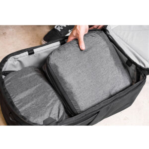 Peak-Design-Packing-Cube-medium--Charcoal6 Henkilö asettaa Peak Design Packing Cube Medium - Charcoal -kuution mustaan matkalaukkuun toisen kuution viereen, ja taustalla näkyy osa kengästä.