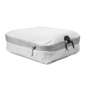 Peak Design Packing Cube Medium - Raw on suorakulmainen valkoinen kuutio, jossa on harmaa vetoketju, osittain täytetty ja hieman pullistunut valkoisella pohjalla. Sen osittain auki oleva vetoketju paljastaa sen kapasiteetin organisoituun pakkaamiseen.
