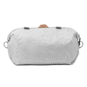 Peak Design Shoe Pouch - Raw on vaaleanharmaa vetoketjullinen pussi, jossa on kuvioitu kuvio, kaksi sivulenkkiä ja pieni ruskea aksenttilappu päällä valkoisella pohjalla.