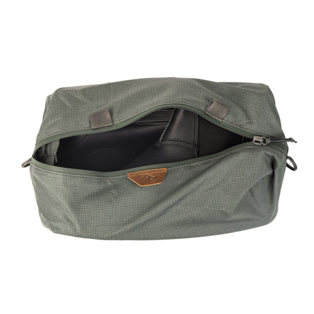 Peak-Design-Shoe-Pouch---Sage3 Peak Design Shoe Pouch, väri Sage, on osittain avattu, ja sen sisältä paljastuu mustat kengät. Pussissa on pieni ruskea nahkalappu edessä ja kangaslenkit sivuilla.
