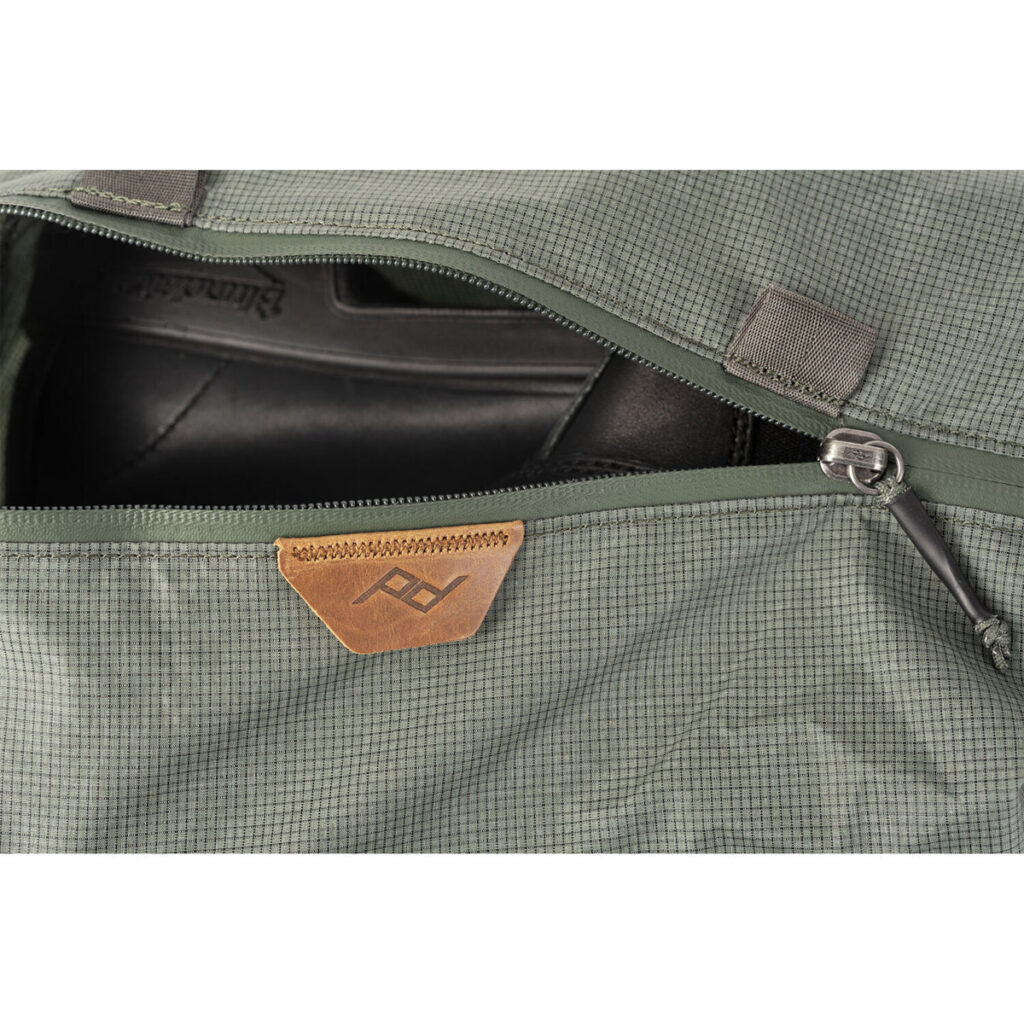 Peak-Design-Shoe-Pouch---Sage4 Lähikuva avoimesta Peak Design Shoe Pouch -kenkäpussista, väri Sage, josta paljastuu osittain mustat kengät. Pussissa on pieni ruskehtava nahkalappu, jossa on geometrinen logo.