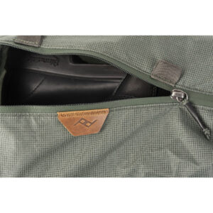 Peak-Design-Shoe-Pouch---Sage4 Lähikuva avoimesta Peak Design Shoe Pouch -kenkäpussista, väri Sage, josta paljastuu osittain mustat kengät. Pussissa on pieni ruskehtava nahkalappu, jossa on geometrinen logo.