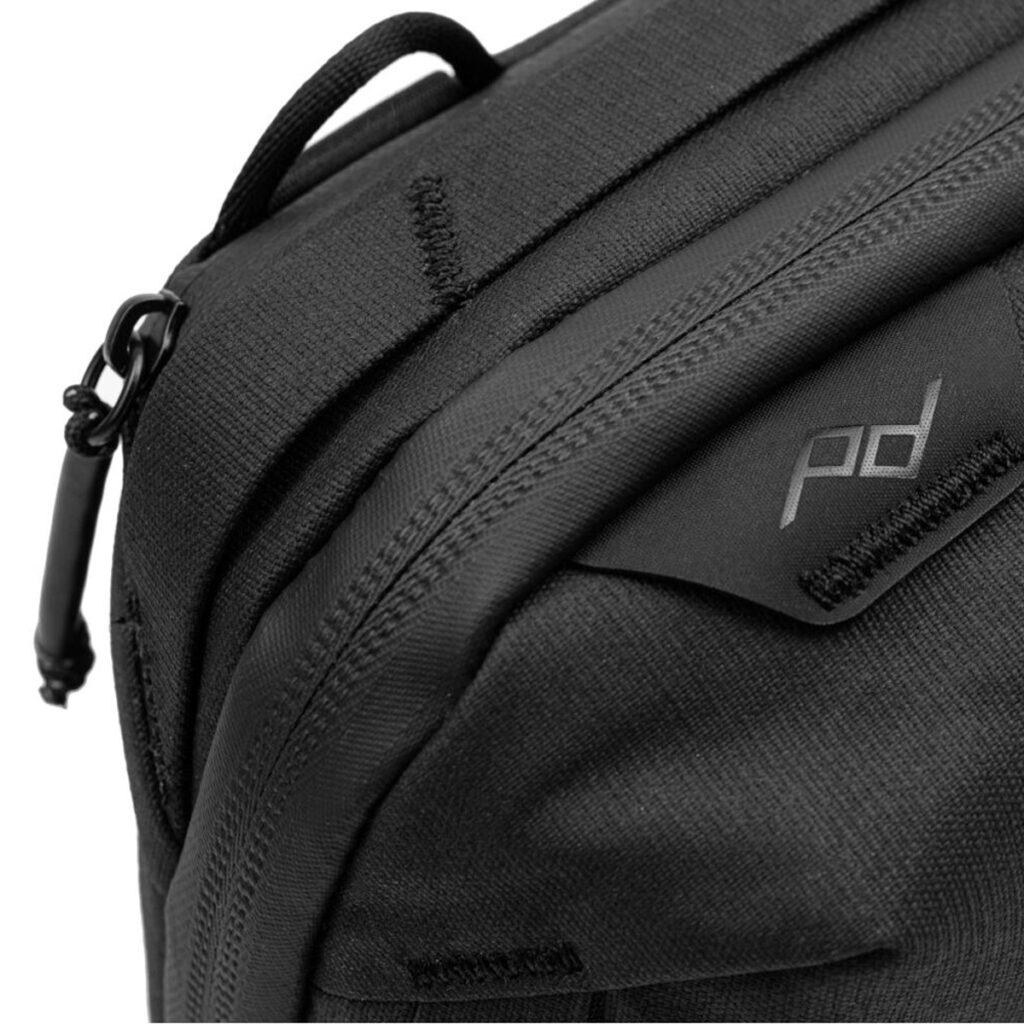 Peak-Design-Tech-Pouch-Black-3 Lähikuva Peak Design Tech Pouch - Black -laukusta, jossa näkyy vetoketju vetokielekkeellä, pieni pd-logolla varustettu lappu sekä näkyvä kankaan rakenne ja ompeleiden yksityiskohdat.