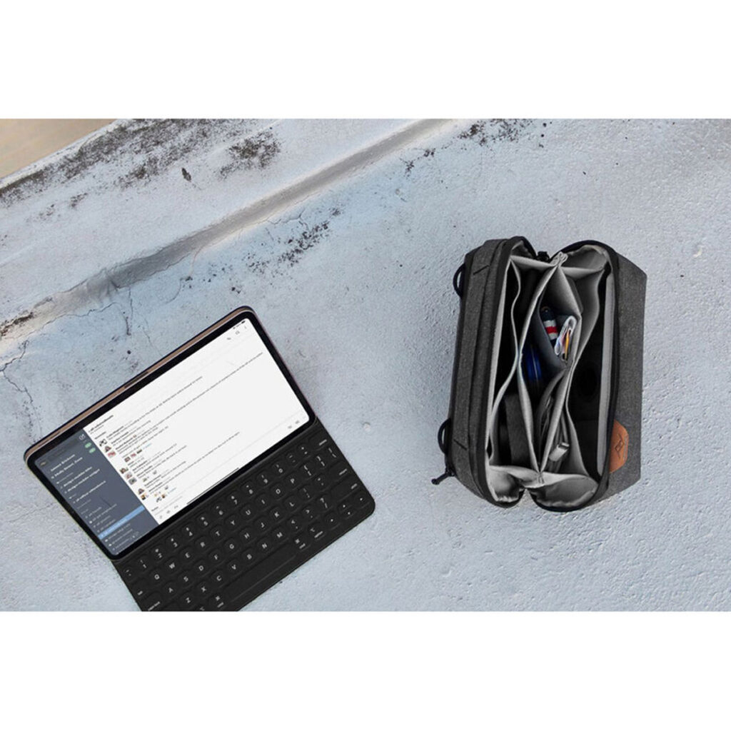 Peak-Design-Tech-Pouch-Black-4 Tabletti, jossa on näppäimistö ja jossa näkyy viestisovellus, istuu betonilla avoimen mustan Peak Design Tech Pouch -laukun vieressä, jossa on johtoja, kyniä ja elektroniikkaa.