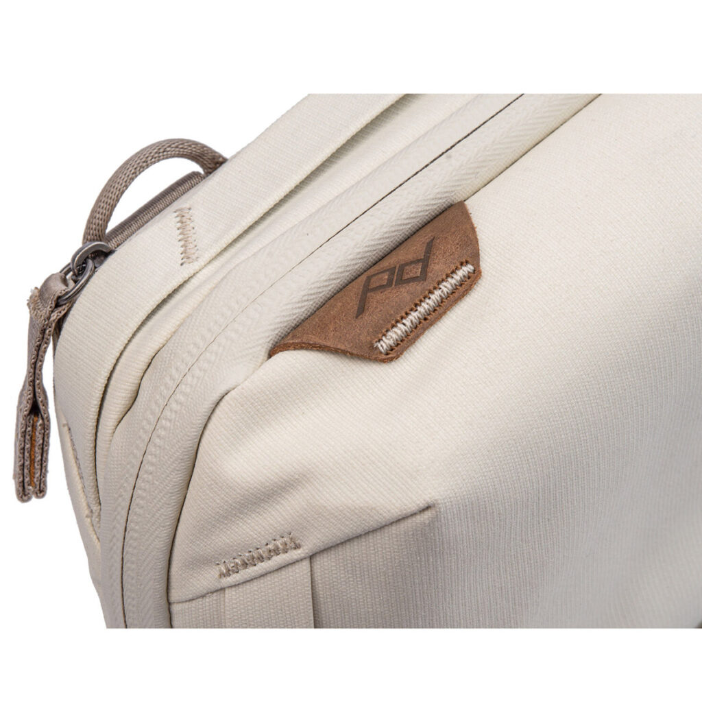 Peak-Design-Tech-Pouch-Bone-3 Lähikuva Peak Design Tech Pouch - Bone -laukusta, joka on beige kangasorganisaattori, jossa on ruskea nahkainen aksentti, jossa on PD-logo, vetoketju, jossa on kangasvetoketju ja yksityiskohtaiset ompeleet.