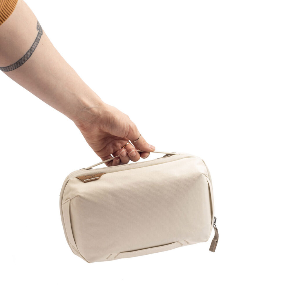 Peak-Design-Tech-Pouch-Bone-4 Henkilön käsi pitelee Peak Design Tech Pouch - Bone -laukkua sen kahvasta valkoista taustaa vasten. Pienessä beigessä pussissa on yksinkertainen, minimalistinen muotoilu.
