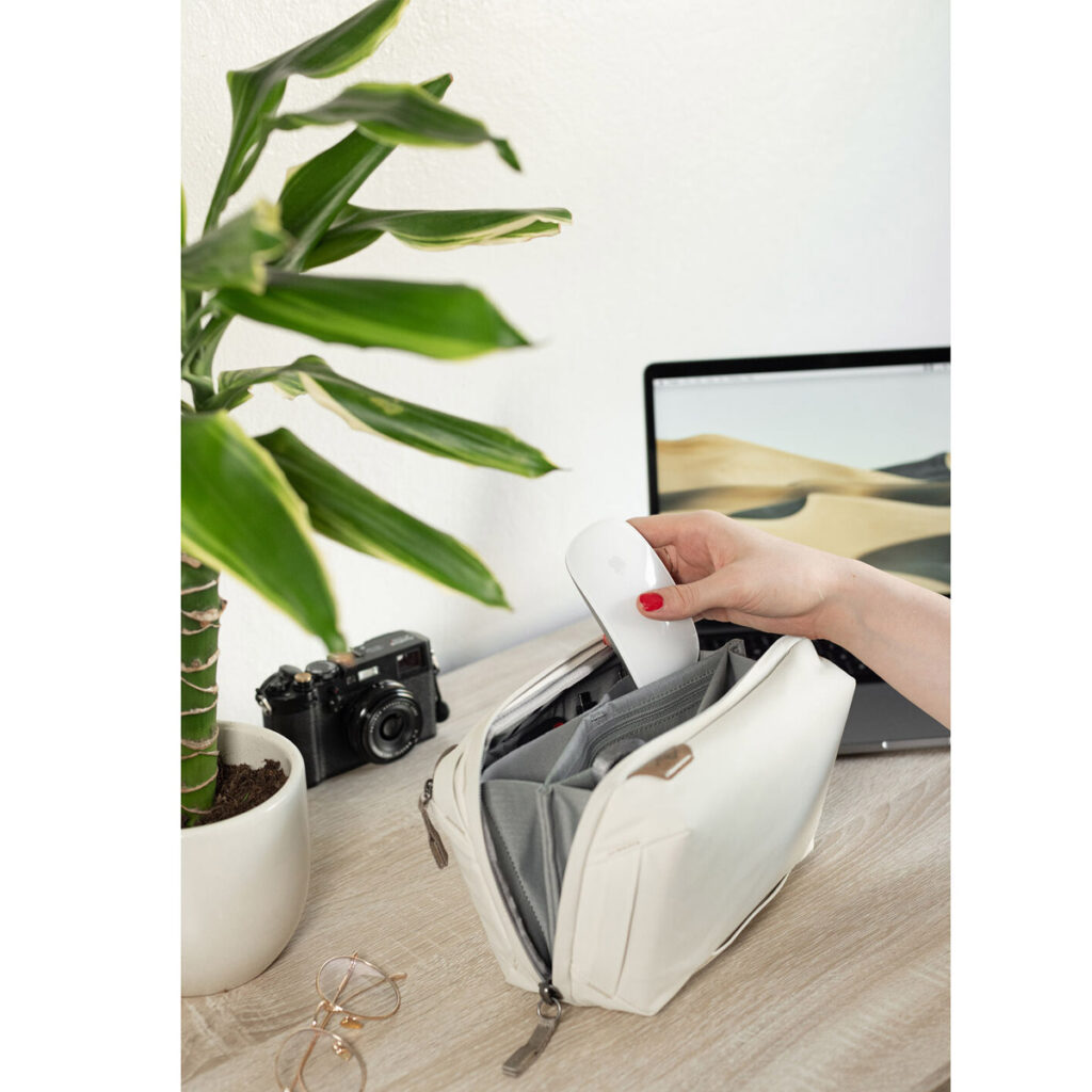 Peak-Design-Tech-Pouch-Bone-6 Käsi laittaa valkoisen langattoman hiiren Peak Design Tech Pouch - Bone -laukkuun pöydällä, jossa on ruukkukasvi, vintage-kamera, silmälasit ja auki oleva kannettava tietokone, jossa näkyy aavikkomaisema.