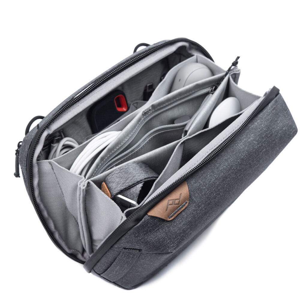 Peak-Design-Tech-Pouch-Charcoal-2 Peak Design Tech Pouch - Charcoal on harmaa organisointilaukku, jossa on useita lokeroita ja johon mahtuu vempaimia ja lisävarusteita, kuten tietokonehiiri, punainen ja musta USB-asema, kaapeleita ja muita pieniä elektroniikkatarvikkeita.
