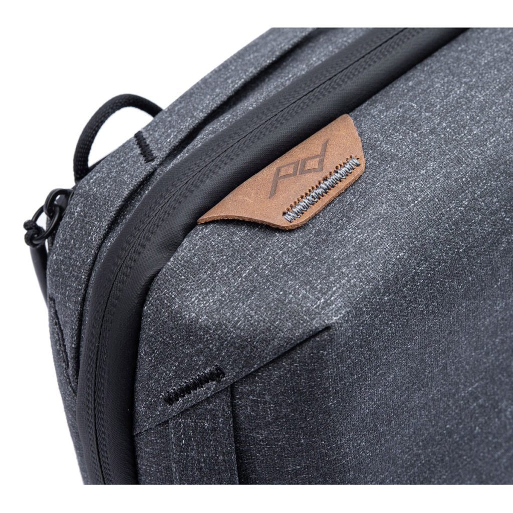 Peak-Design-Tech-Pouch-Charcoal-3 Lähikuva Peak Design Tech Pouch -laukusta, jossa on harmaa kangas, musta vetoketju ja ruskea nahkainen välilehti, johon on kohokuvattu "pd". Pussissa on moderni, minimalistinen muotoilu.