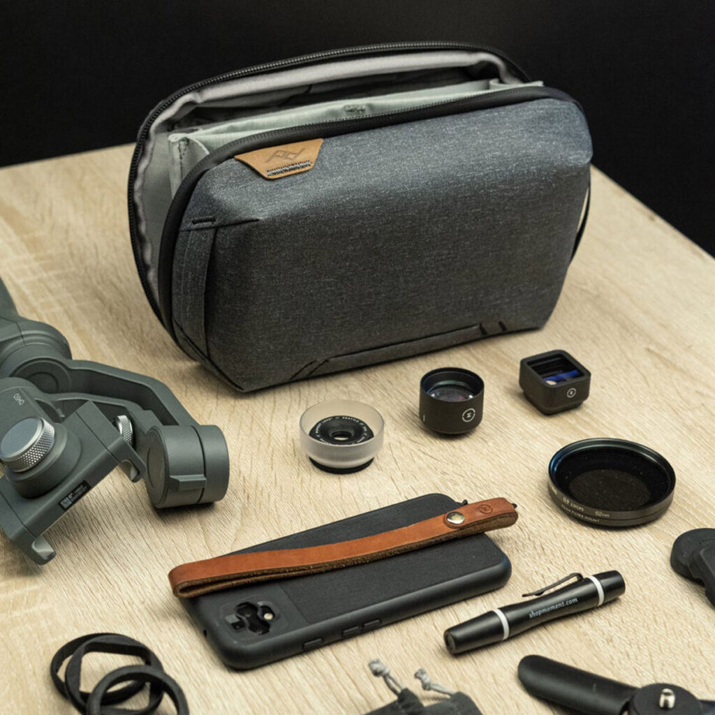 Peak-Design-Tech-Pouch-Charcoal-4 Peak Design Tech Pouch - Charcoal lepää puisella pöydällä, siististi kameratarvikkeiden, puhelimen objektiivien, nahkahihnalla varustetun älypuhelimen, pienen taskulampun, kardaanin ja linssinsuojusten ympäröimänä.