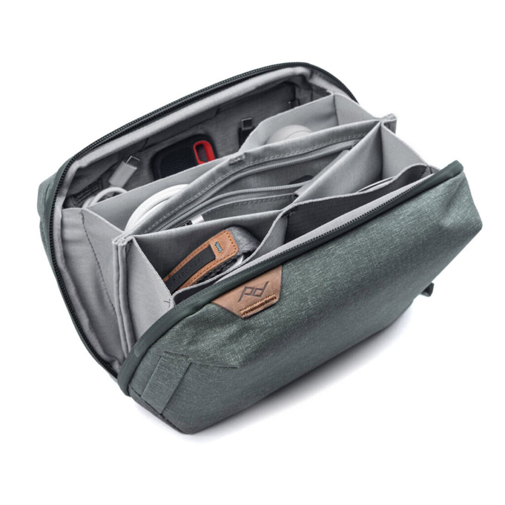 Peak-Design-Tech-Pouch-Sage-2 Peak Design Tech Pouch - Sage on osittain auki ja paljastaa järjestetyn säilytystilan tarvikkeille useissa pehmustetuissa lokeroissa ja väliseinissä, joiden etupuolella on näkyvissä nahkalaikka, jossa on logo.
