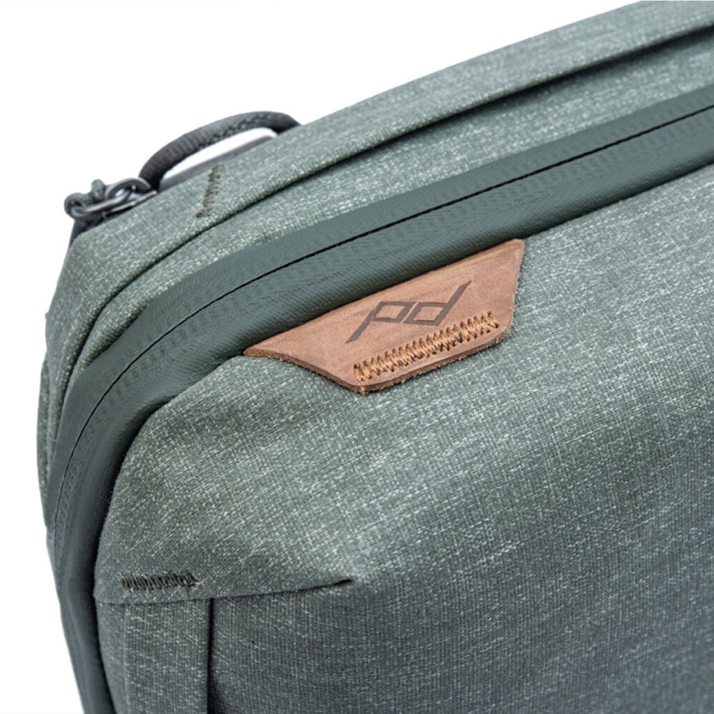 Peak-Design-Tech-Pouch-Sage-3 Lähikuva Peak Design Tech Pouch -laukusta värissä Sage, jossa on vihreä kangas, jossa on ruskea nahka-aksentti, pd-logo, ylänurkassa oleva kahva ja vetoketju.