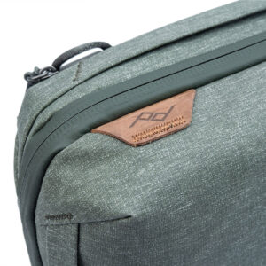 Peak-Design-Tech-Pouch-Sage-3 Lähikuva Peak Design Tech Pouch -laukusta värissä Sage, jossa on vihreä kangas, jossa on ruskea nahka-aksentti, pd-logo, ylänurkassa oleva kahva ja vetoketju.