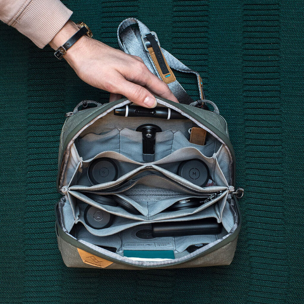 Peak-Design-Tech-Pouch-Sage-4 Käsi pitää auki Peak Design Tech Pouch - Sage -laukkua, jonka sisältä paljastuu siististi järjestettyjä kameran linssejä, tarvikkeita ja laitteita, jotka ovat kaikki aseteltuina kuvioidulle vihreälle pinnalle.