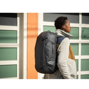 Peak-Design-Travel-Backpack-30L-black7 Ulkona oleva henkilö seisoo sivuttain rakennuksen edessä, hänellä on yllään harmaa ja ruskea fleecetakki ja hänellä on selässään musta Peak Design Travel Backpack 30L -kamerareppu.
