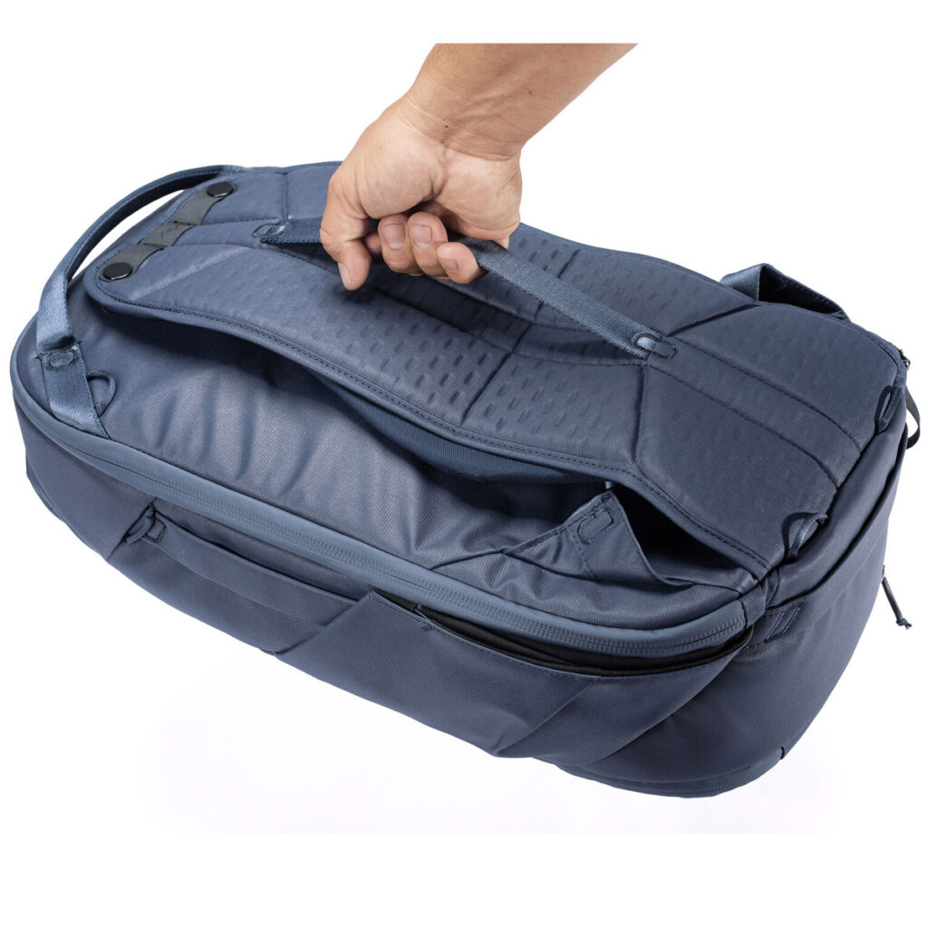 Peak-Design-Travel-Backpack-30L-midnight6 Käsi pitää Peak Design Travel Backpack 30L -kamerareppu, tummansininen sen yläkahvasta, jolloin laivastonsinisen laukun sivu- ja vetoketjulliset lokerot näkyvät valkoista taustaa vasten.