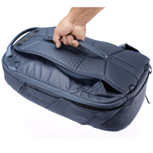 Peak-Design-Travel-Backpack-30L-midnight6 Käsi pitää Peak Design Travel Backpack 30L -kamerareppu, tummansininen sen yläkahvasta, jolloin laivastonsinisen laukun sivu- ja vetoketjulliset lokerot näkyvät valkoista taustaa vasten.