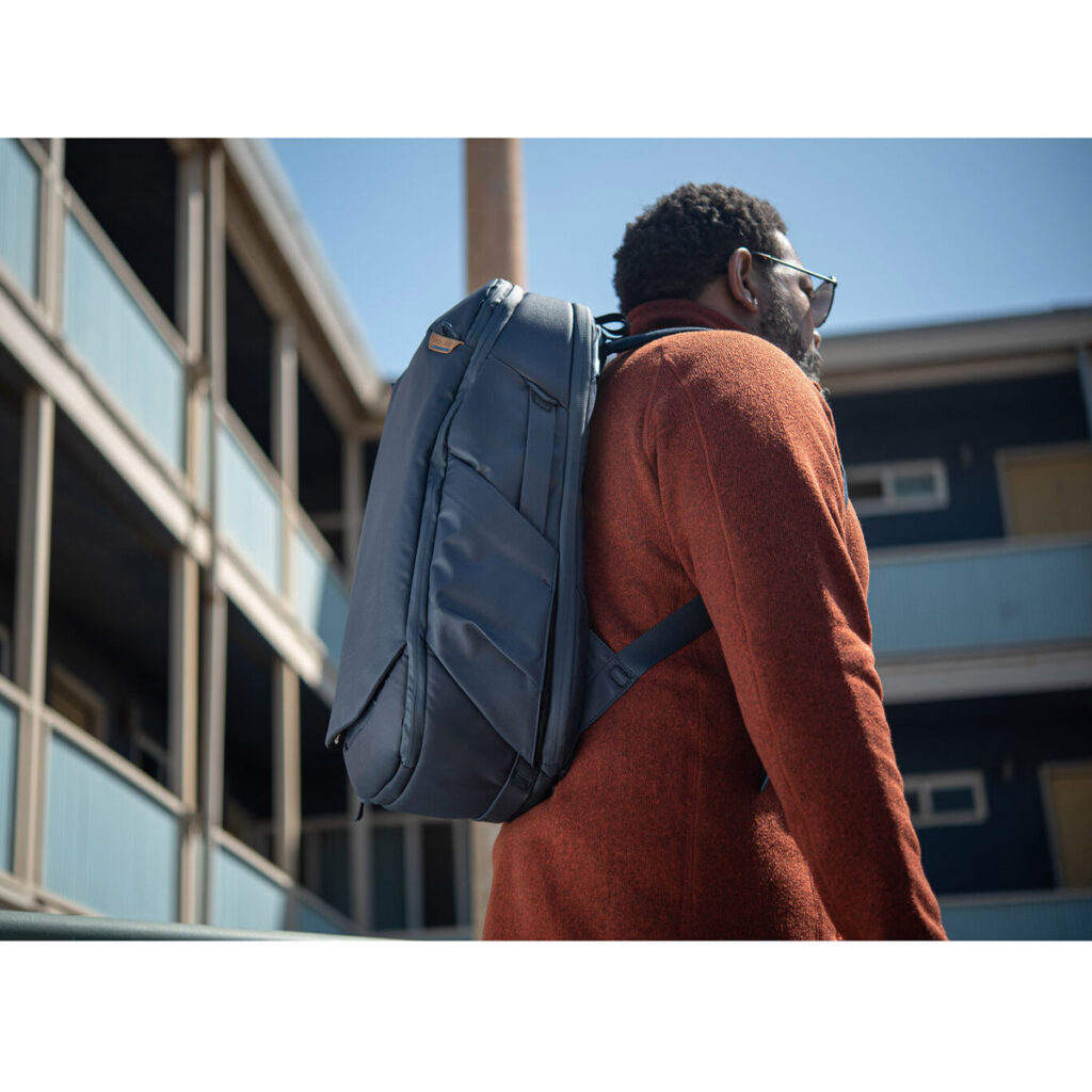 Peak-Design-Travel-Backpack-30L-midnight7 Aurinkolaseihin ja oranssiin takkiin pukeutunut mies seisoo ulkona Peak Design Travel Backpack 30L -kamerareppu, tummansininen. Hänen takanaan on monikerroksinen rakennus, jossa on siniset kaiteet ja savupiippu.