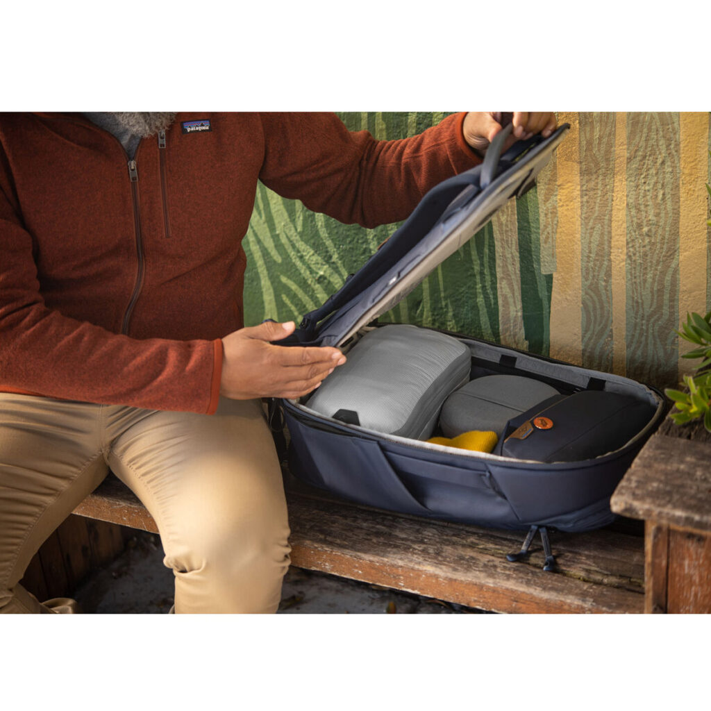 Peak-Design-Travel-Backpack-30L-midnight8 Punatakkinen ja beigeihin housuihin pukeutunut henkilö istuu puisella penkillä ja avaa Peak Design Travel Backpack 30L -kamerareppu, tummansininen, jonka sisällä on siististi pakattuja tavaroita ja jonka takana on lehtivihreä ja keltainen taustaseinä.