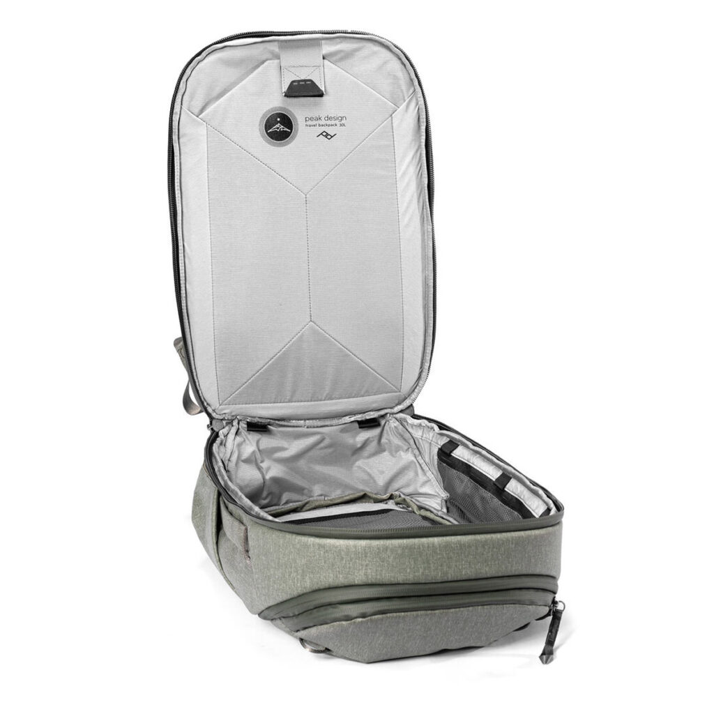Peak-Design-Travel-Backpack-30L-sage4 Peak Design Travel Backpack 30L -kamerareppu, vaaleanvihreä, seisoo pystyasennossa, kun sen päälokero on avattu vetoketjuineen, jolloin paljastuu vaaleanharmaa sisustus, jossa on taskuja ja järjestysosioita. Kansi nojaa taaksepäin, jotta siihen pääsee helposti käsiksi.