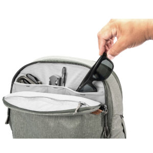 Peak-Design-Travel-Backpack-30L-sage5 Käsi asettaa mustat aurinkolasit vaaleanvihreä Peak Design Travel Backpack 30L -kamerareppu, johon mahtuu myös monitoimityökalu, kynä ja muita tavaroita, järjestettyyn taskuun avoimessa päälokerossa.