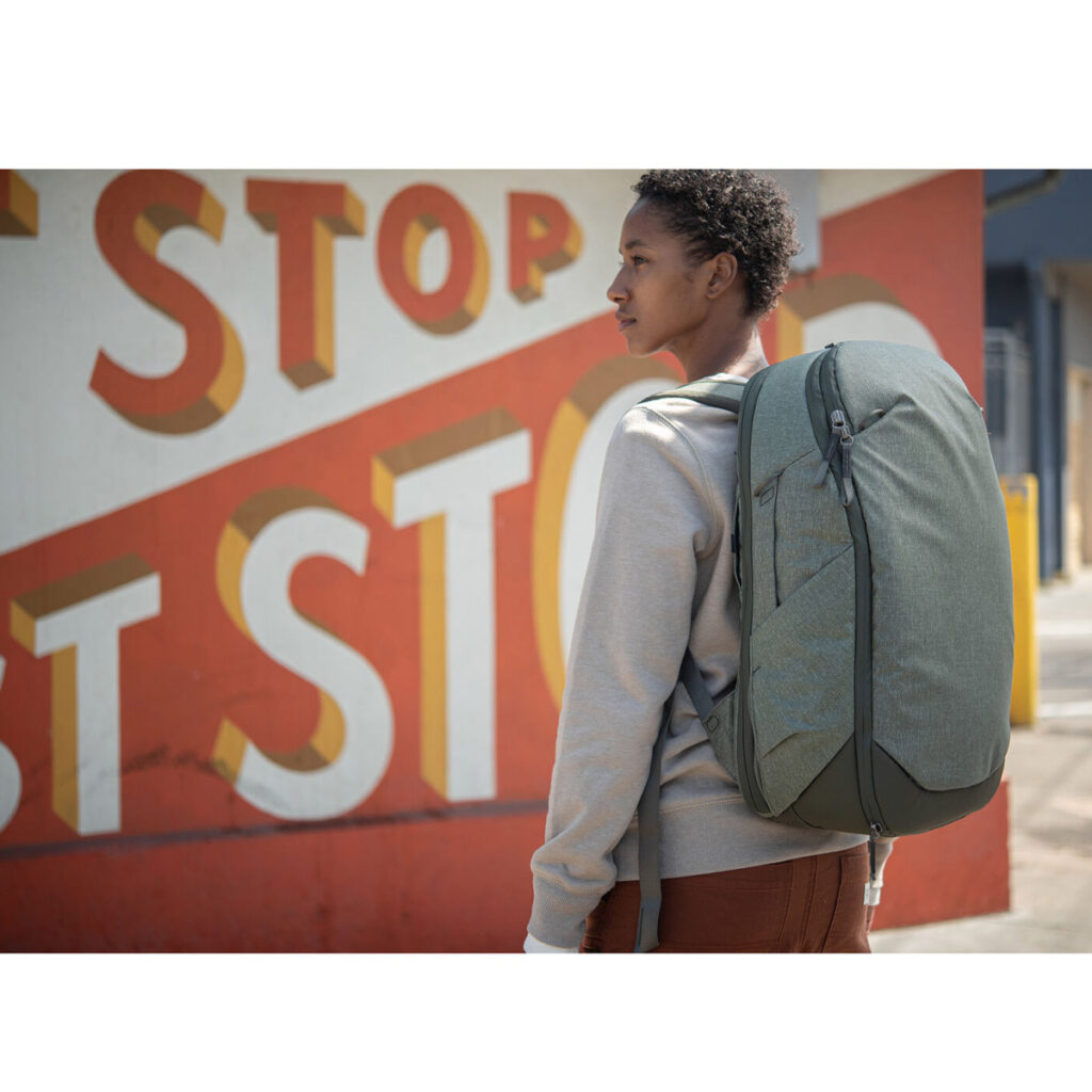 Peak-Design-Travel-Backpack-30L-sage7 Henkilö, jolla on yllään Peak Design Travel Backpack 30L -kamerareppu, vaaleanvihreä, seisoo ulkona värikkään seinämaalauksen edessä, jossa on suuria punaisia ja keltaisia kirjaimia, joissa lukee STOP.