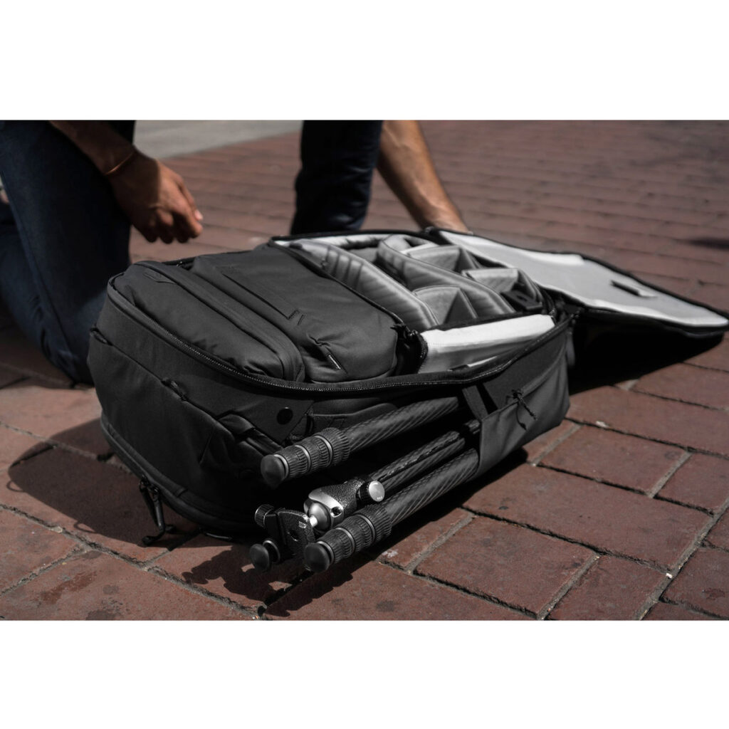 Peak-Design-Travel-Backpack-45L-black09 Henkilö polvistuu tiilipäällysteisellä jalkakäytävällä avoimen Peak Design Travel Backpack 45L -kamerareppu, musta, jonka sisälle mahtuu kameravarusteet ja sen ulkolokeroon osittain säilytetty jalusta.