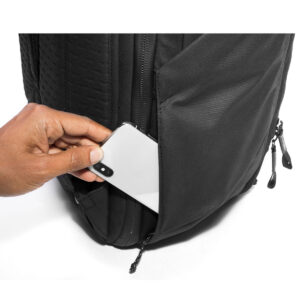 Peak-Design-Travel-Backpack-45L-black10 Käsi laittaa valkoisen älypuhelimen mustan Peak Design Travel Backpack 45L -kamerareppu -repun vetoketjulliseen sivutaskuun, jossa on pääosin valkoinen tausta.