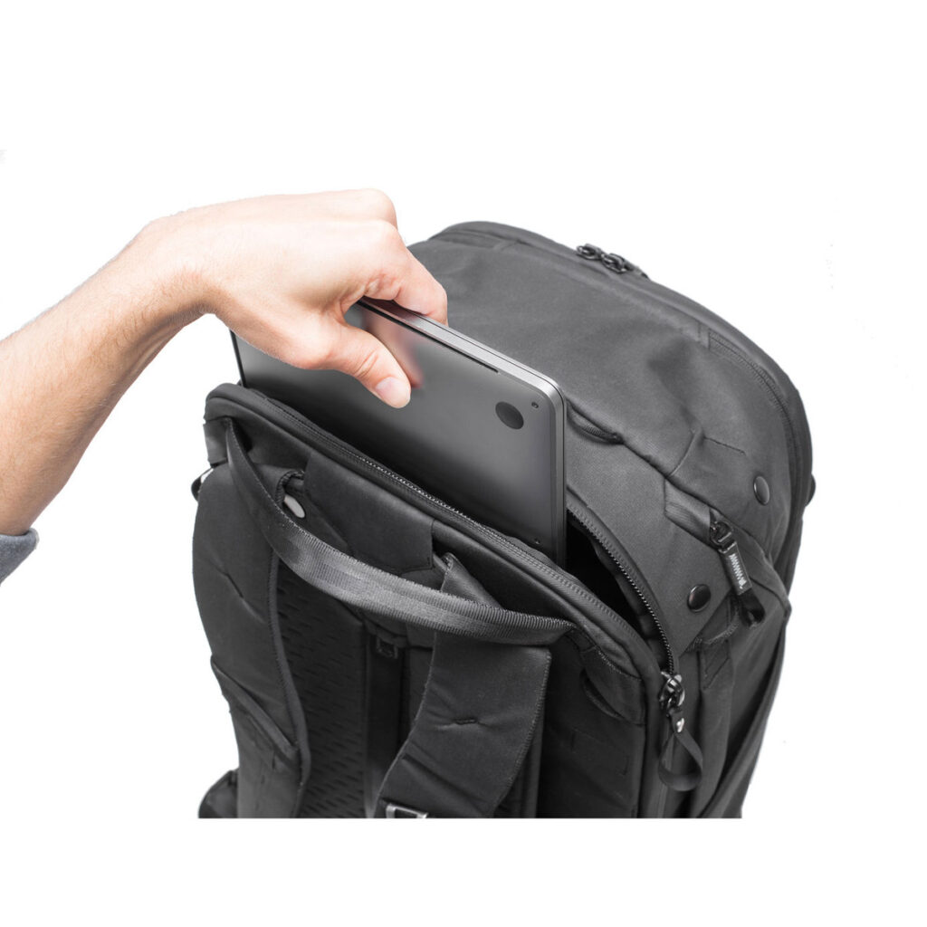Peak-Design-Travel-Backpack-45L-black11 Käsi asettaa kannettavan tietokoneen mustan Peak Design Travel Backpack 45L -kamerareppu -repun etulokeroon valkoista taustaa vasten.