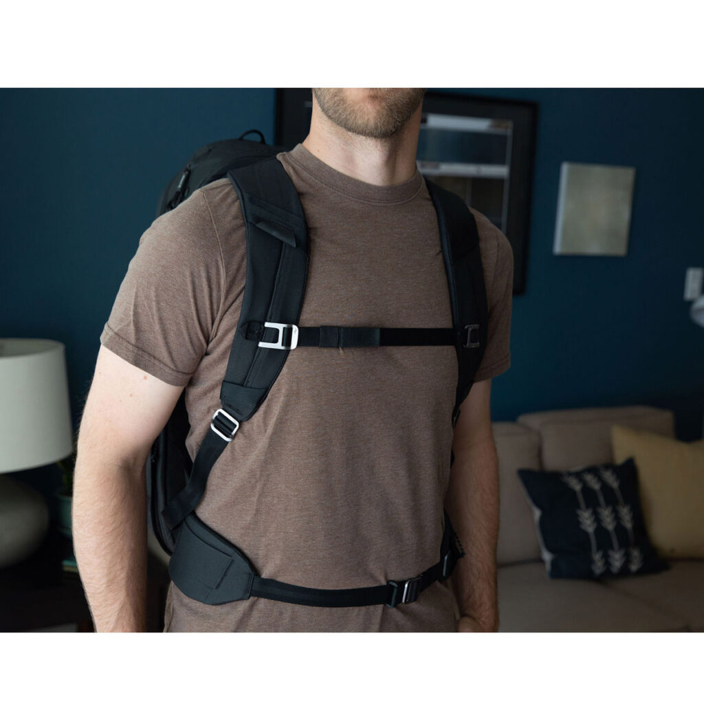 Peak-Design-Travel-Backpack-45L-black12 Ruskeaan t-paitaan pukeutunut henkilö, jolla on mukanaan Peak Design Travel Backpack 45L -kamerareppu, musta seisoo olohuoneessa, jossa on sohva, tyynyjä, pöytävalaisin ja seinätaidetta. Hänen kasvonsa ovat osittain kuvan ulkopuolella.