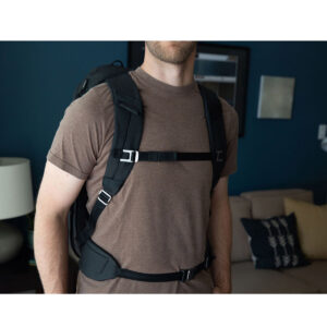 Peak-Design-Travel-Backpack-45L-black12 Ruskeaan t-paitaan pukeutunut henkilö, jolla on mukanaan Peak Design Travel Backpack 45L -kamerareppu, musta seisoo olohuoneessa, jossa on sohva, tyynyjä, pöytävalaisin ja seinätaidetta. Hänen kasvonsa ovat osittain kuvan ulkopuolella.