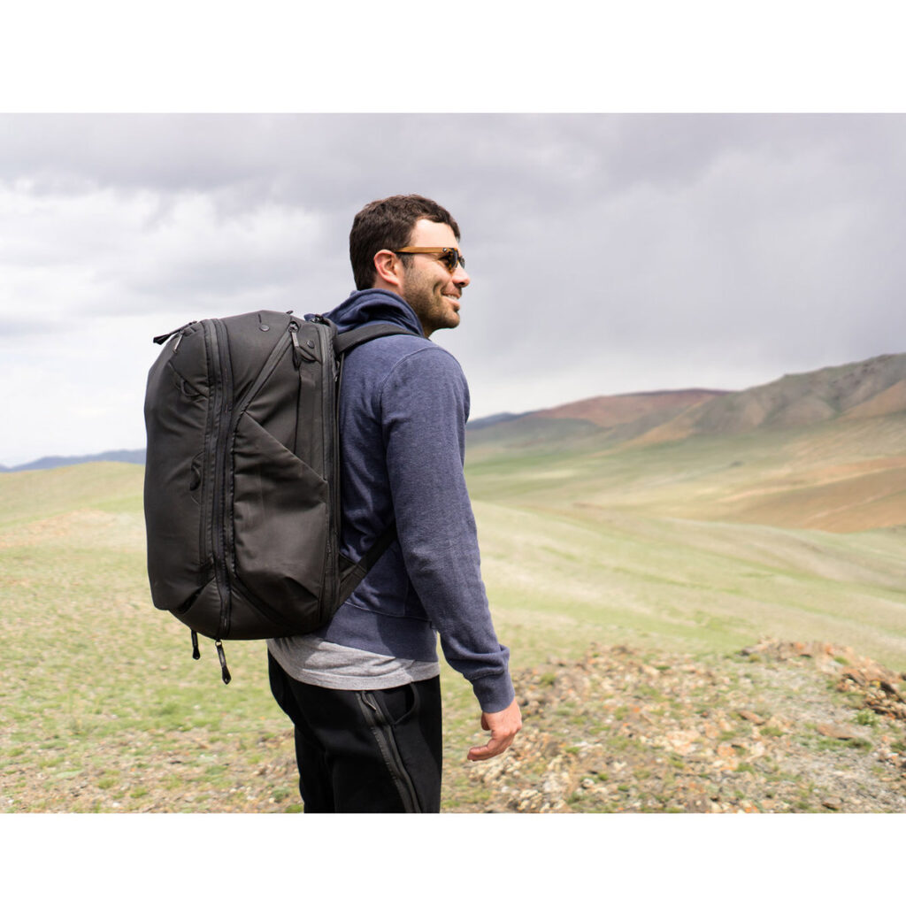 Peak-Design-Travel-Backpack-45L-black13 Aurinkolaseihin ja siniseen huppariin pukeutunut mies seisoo ruohoisella kukkulalla, jossa on pilvinen taivas ja kaukaiset vuoret, ja hänellä on Peak Design Travel Backpack 45L -kamerareppu, musta.