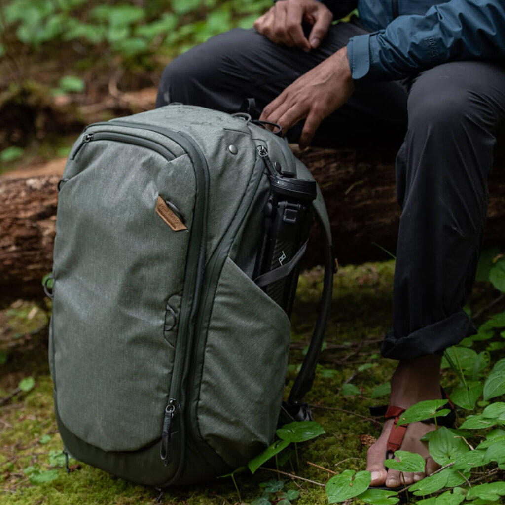 Peak-Design-Travel-Backpack-45L-sage2 Henkilö istuu kaatuneella tukilla metsässä ja säätää viereensä asetettua Peak Design Travel Backpack 45L -kamerareppua, vaaleanvihreä. Hänellä on tummat housut, sininen takki ja punaiset sandaalit vihreällä lehtipohjalla.