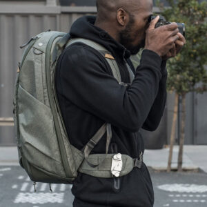 Peak-Design-Travel-Backpack-45L-sage3 Henkilö, jolla on yllään vaaleanvihreä Peak Design Travel Backpack 45L -kamerareppu ja musta huppari, seisoo ulkona, pitää kameraa kasvojaan vasten ja ottaa valokuvaa.