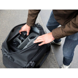 Peak-Design-Travel-Duffel-35l-black-7 Ruskeaan takkiin ja farkkuihin pukeutunut henkilö järjestelee Peak Design Travel Duffel 35L - Black -reppua ja laittaa kameran sisään objektiivien ja tarvikkeiden viereen. Avattu laukku paljastaa sen pehmustetun sisätilan, joka on suunniteltu kameravarusteille.