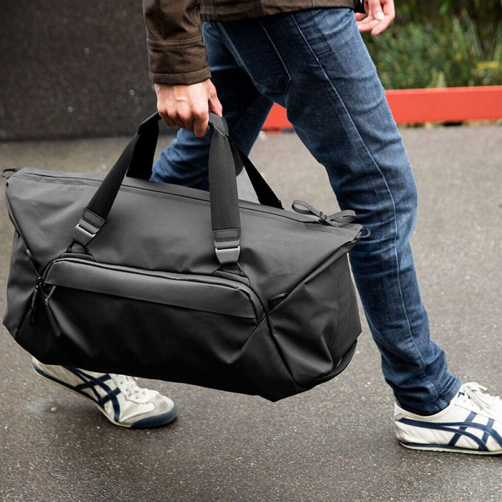 Peak-Design-Travel-Duffel-35l-black-8 Farkkuihin ja lenkkareihin pukeutunut henkilö kävelee jalkakäytävällä ja kantaa Peak Design Travel Duffel 35L - Black -mallistoa kahvoista.