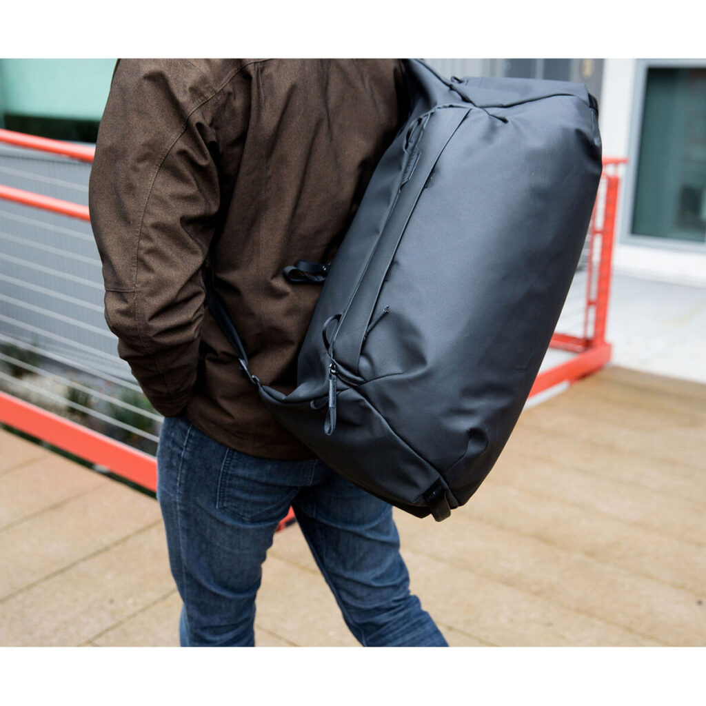 Peak-Design-Travel-Duffel-35l-black-9 Ruskeaan takkiin ja sinisiin farkkuihin pukeutunut henkilö kantaa olkapäänsä yli Peak Design Travel Duffel 35L - Black -reppua kävellessään ulkona lähellä punaista kaidetta.