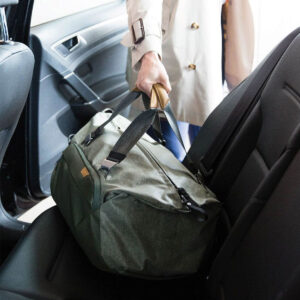 Peak-Design-Travel-Duffel-35l-sage-7 Henkilö, jolla on yllään kevyt trenssitakki, asettaa Peak Design Travel Duffel 35L - Sage -matkakassin mustilla hihnoilla auton takapenkille, jossa on avoin ovi ja musta sisustus.