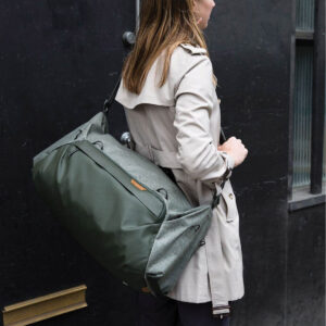 Peak-Design-Travel-Duffel-35l-sage-8 Beigeen trenssitakkiin pukeutunut nainen seisoo tumman seinän vieressä, ja hänellä on olallaan Peak Design Travel Duffel 35L - Sage.