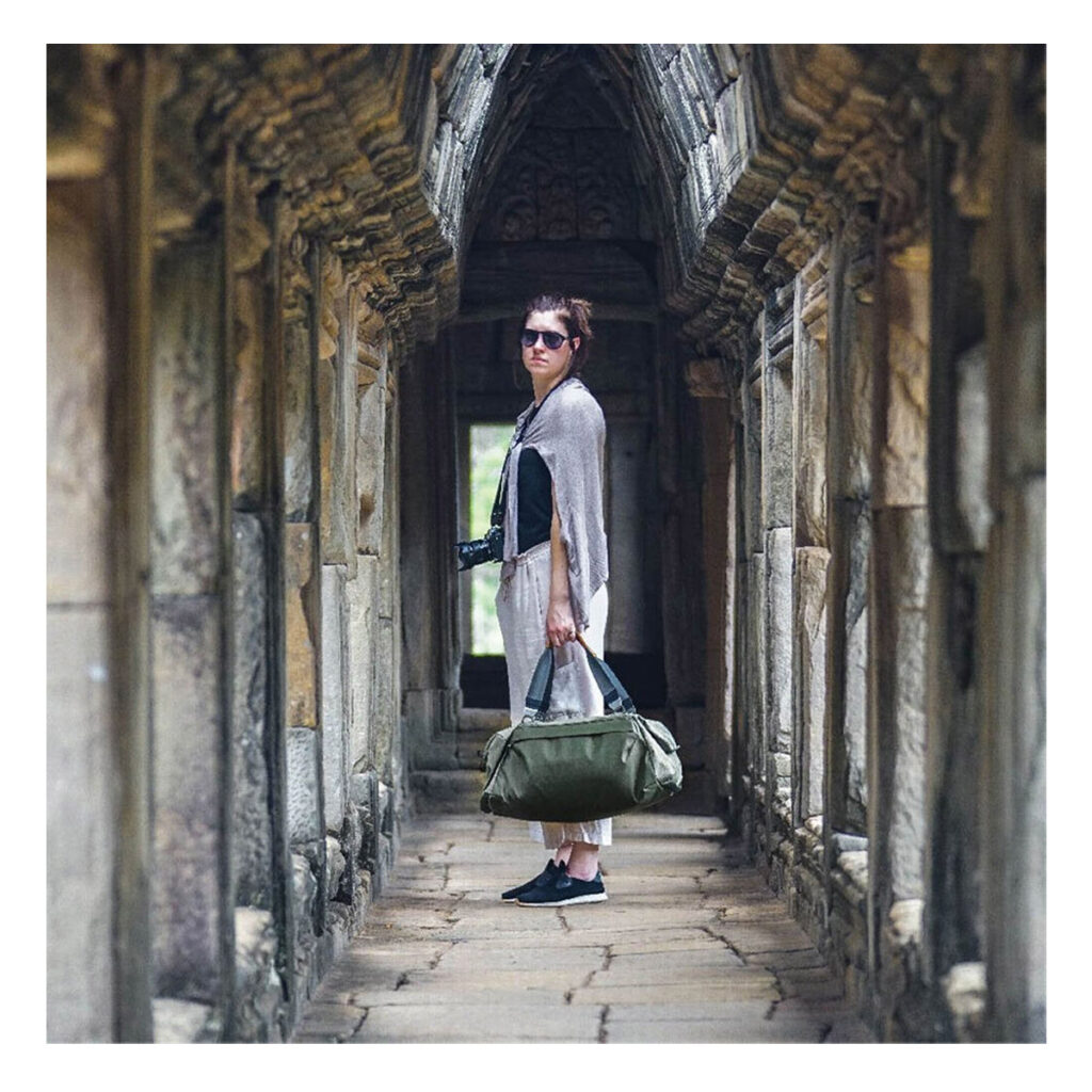 Peak-Design-Travel-Duffel-35l-sage-9 Aurinkolaseihin ja rentoihin vaatteisiin pukeutunut nainen seisoo muinaisessa kivikäytävässä, kädessään Peak Design Travel Duffel 35L - Sage ja kamera, jota ympäröivät veistetyt pylväät ja kaaret.