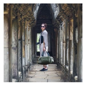 Peak-Design-Travel-Duffel-35l-sage-9 Aurinkolaseihin ja rentoihin vaatteisiin pukeutunut nainen seisoo muinaisessa kivikäytävässä, kädessään Peak Design Travel Duffel 35L - Sage ja kamera, jota ympäröivät veistetyt pylväät ja kaaret.