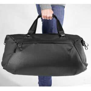 Peak-Design-Travel-Duffel-65l-black-6 Sinisiin farkkuihin ja harmaaseen toppiin pukeutunut henkilö pitää Peak Design Travel Duffel 65L - Black -matkakassia kahvoista käsin valkoista taustaa vasten.