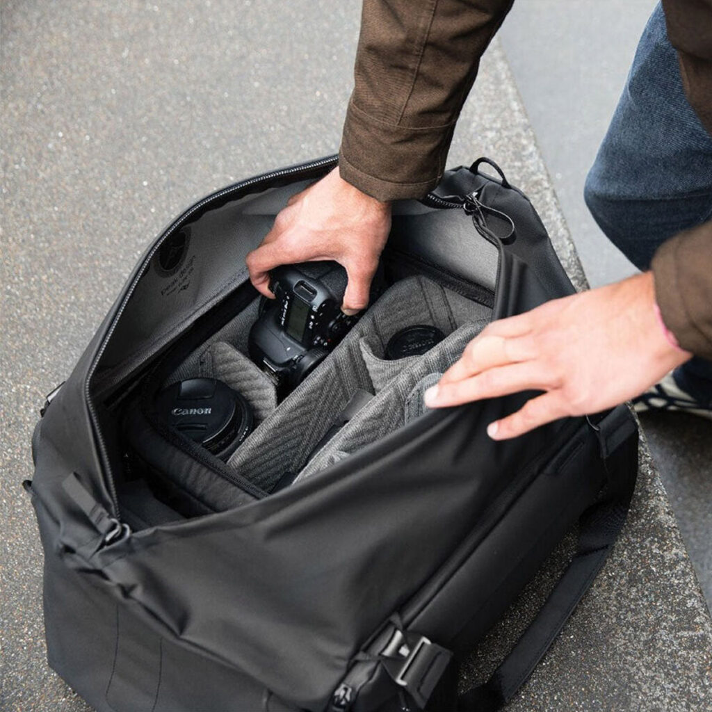 Peak-Design-Travel-Duffel-65l-black-7 Henkilö asettaa Canon-kameran, ylimääräisiä objektiiveja ja tarvikkeita mustaan Peak Design Travel Duffel 65L -matkalaukkuun, jossa on järjestetyt lokerot, ja asettaa sen betonipinnalle.